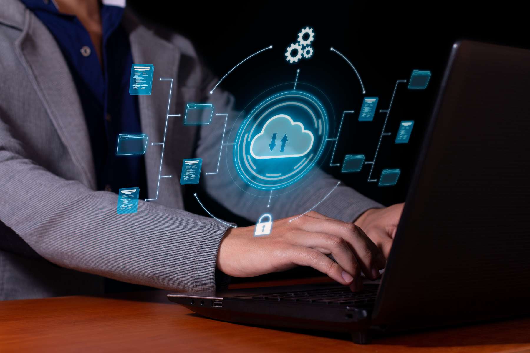 Ein Mitarbeiter im Sakko arbeitet an einem schwarzen Laptop während vor ihm eine digitale Darstellung einer hellblauen Cloud zu sehen ist.