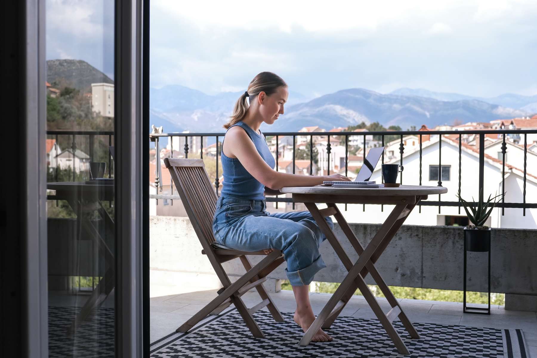 Eine junge Frau in Jeans und Tanktop sitzt auf dem Balkon an einem runden Holztisch auf dem ein Block liegt und eine Kaffeetasse steht und arbeitet an einem Laptop.