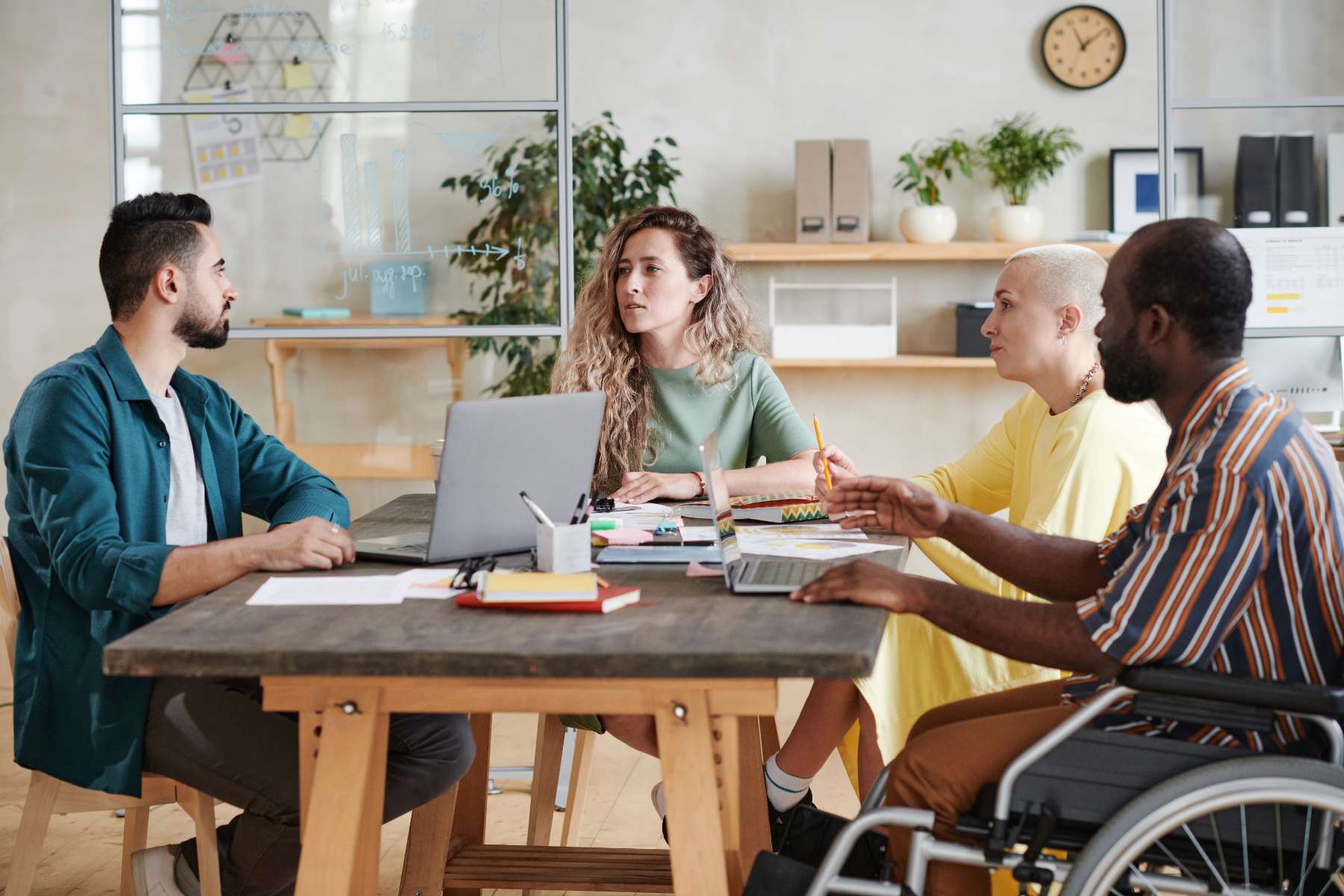 Vier Angestellte, darunter ein Mann im Rollstuhl, sitzen an einem Holztisch auf dem Arbeitsutensilien wie Blätter, Stifte und Laptops liegen und unterhalten sich.