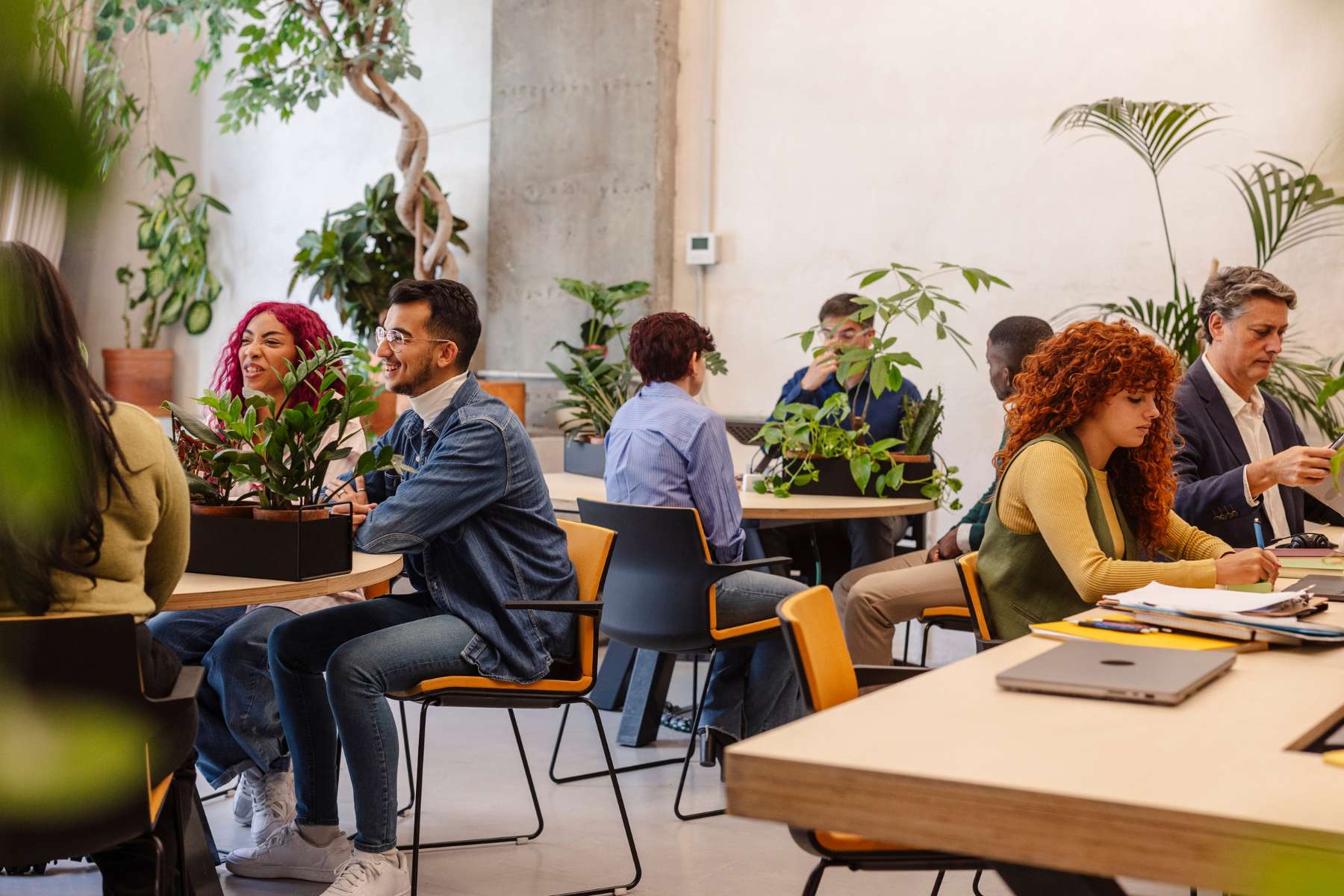 Eine Gruppe diverser Büromitarbeiter sitzt in einem gemütlich eingerichteten Loungebereich an mehreren Tischen aus Holz auf denen Pflanzen stehen, unterhalten sich miteinander oder arbeiten am Laptop.