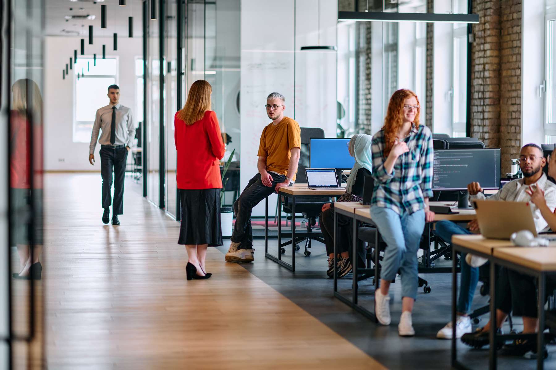 Mehrere junge diverse Angestellte sitzen bzw. stehen an Schreibtischen in einem weiträumigen Büro und unterhalten sich.