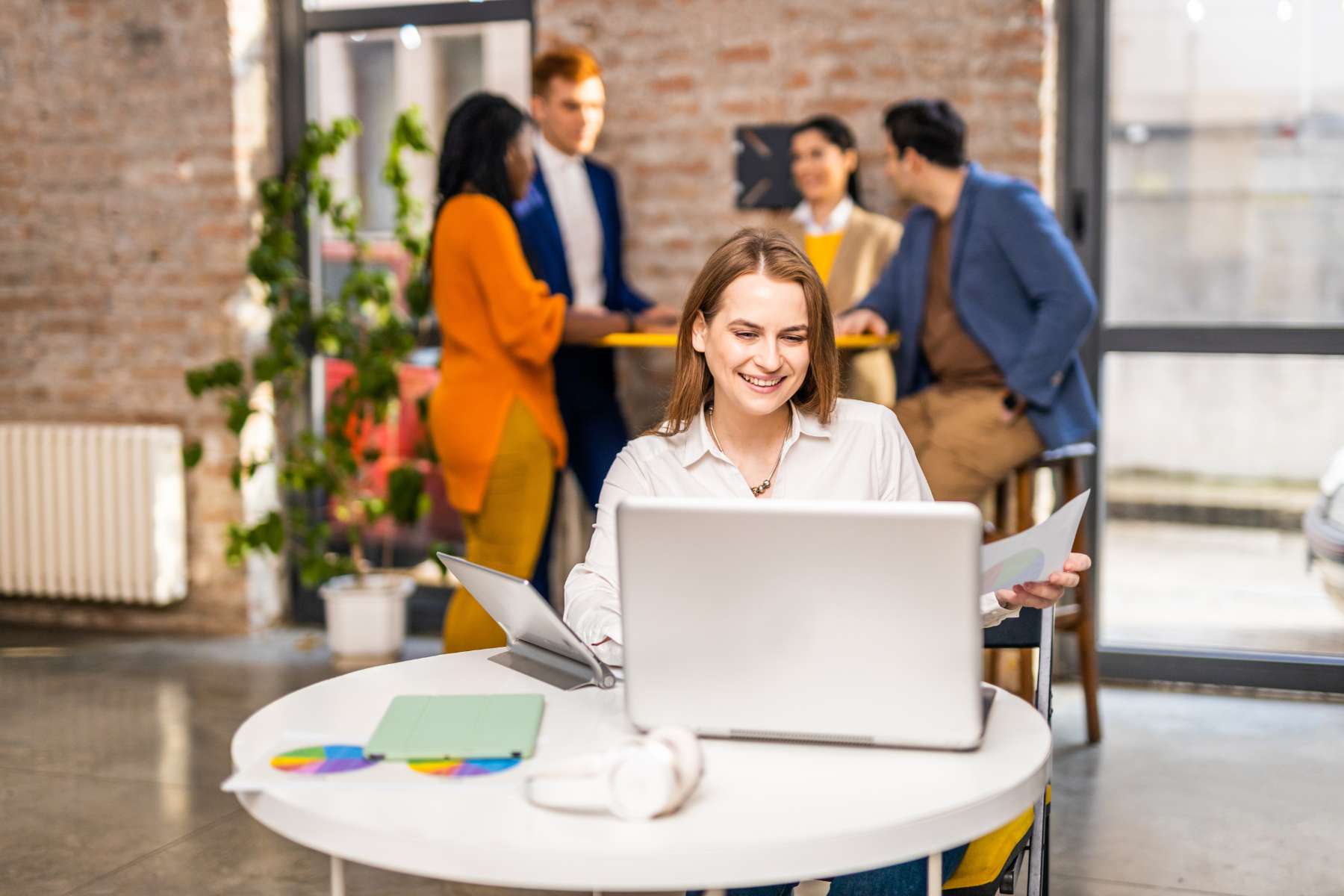 Eine lächelnde Büroangestellte sitzt an einem Tisch vor einem Laptop während sich im Hintergrund 4 Arbeitskollegen an einem Stehtisch unterhalten.