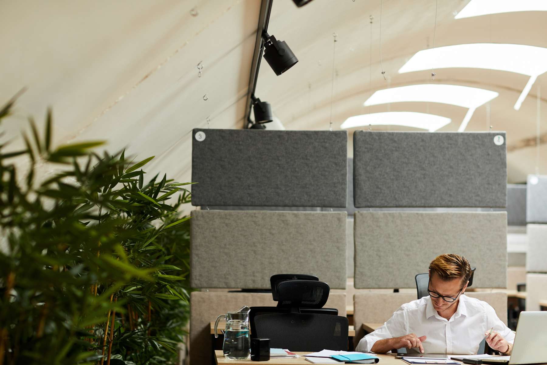 Ein männlicher Angestellter sitzt im Büro an einem Schreibtisch vor einem großen Akustikpaneel und arbeitet am Tablet.