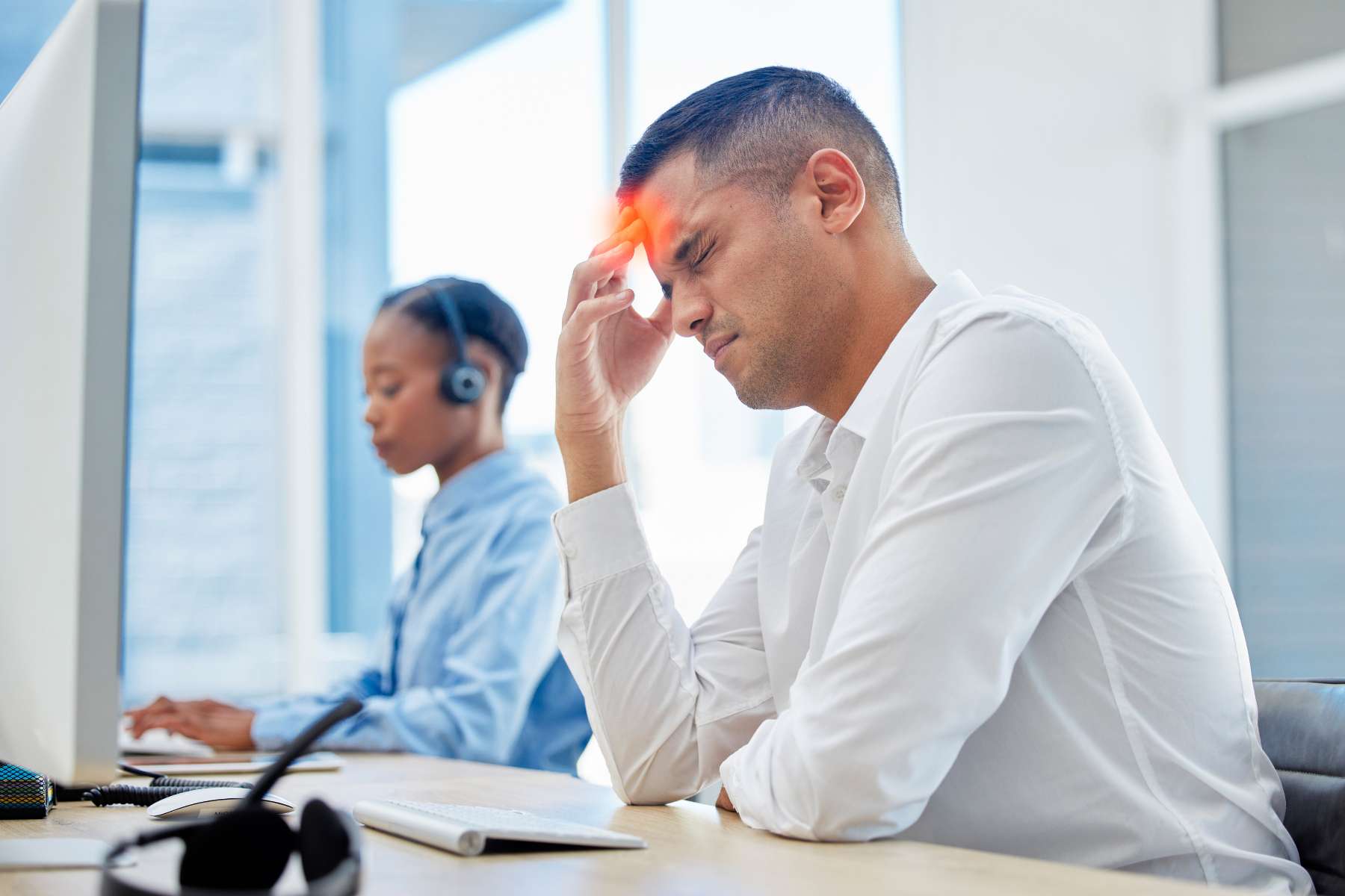 Ein junger Büroangestellter sitzt an einem Schreibtisch vor einem Bildschirm und hält sich mit schmerverzerrtem Gesicht eine Hand an die Stirn, während im Hintergrund eine weibliche Kollegin mit Headset ebenfalls am Laptop arbeitet.
