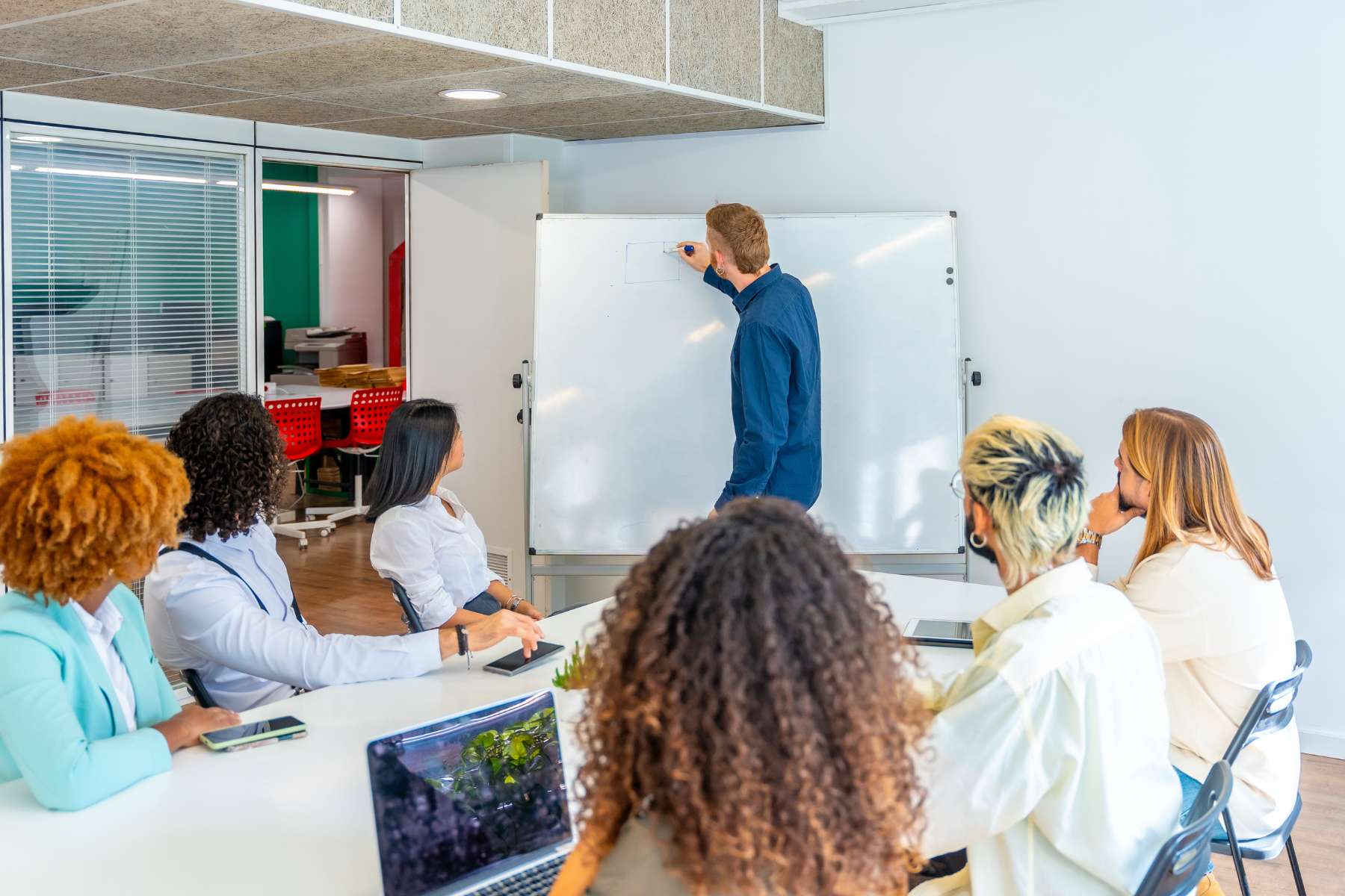 Eine Gruppe junger Büroangestellter sitzt an einem großen weißen Tisch und schaut einem jungen Mann zu, der etwas auf ein Whiteboard schreibt.