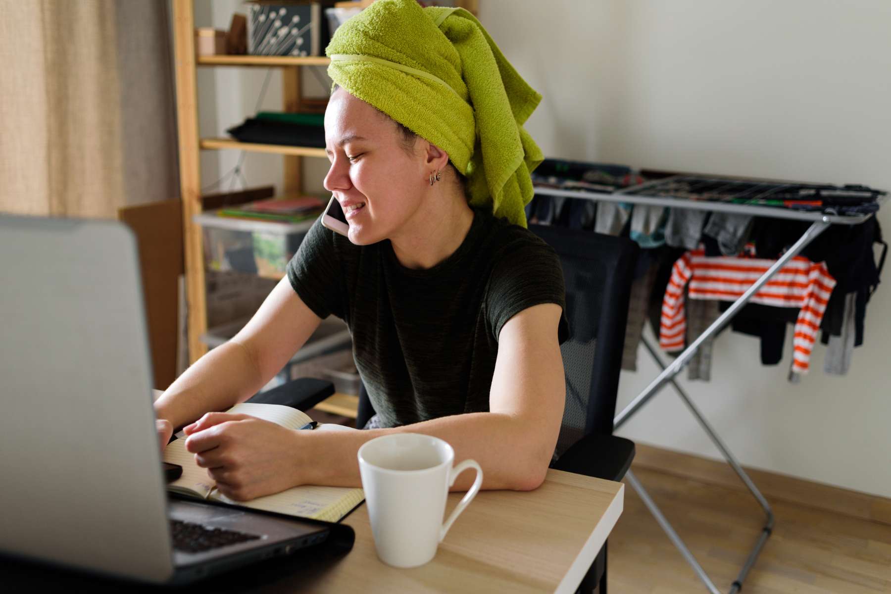 Junge Frau mit Handtuch auf dem Kopf ist im Homeoffice, sitzt an einem Schreibtisch, hat eine Tasse Kaffee neben sich und arbeitet am Laptop während im Hintergrund ein Wäschekorb steht.