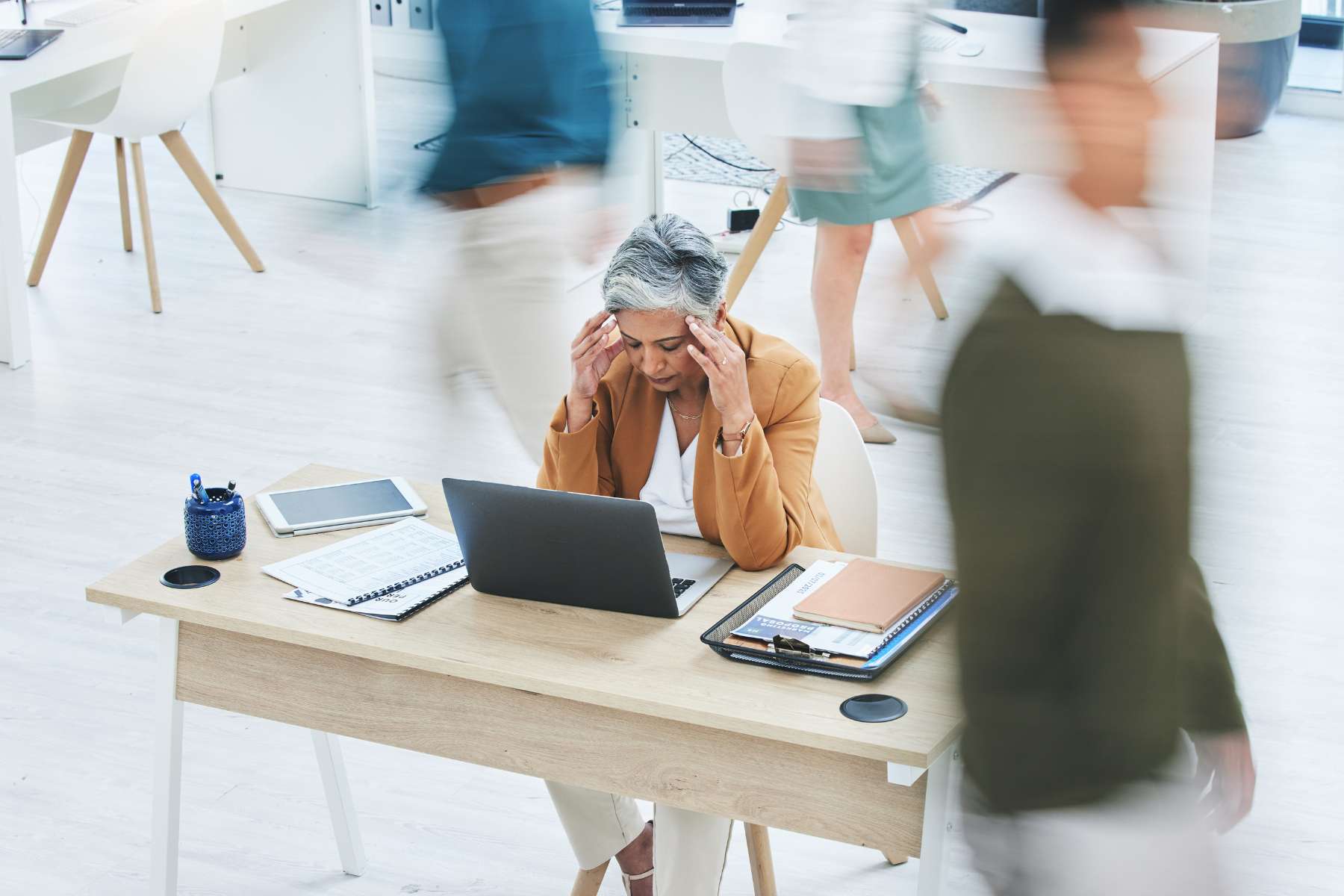 Eine gestresste Büromitarbeiterin sitzt an einem Schreibtisch aus Holz vor einem Laptop und hält beide Hände an die Schläfen, während um sie herum im Büro alles in Bewegung ist.