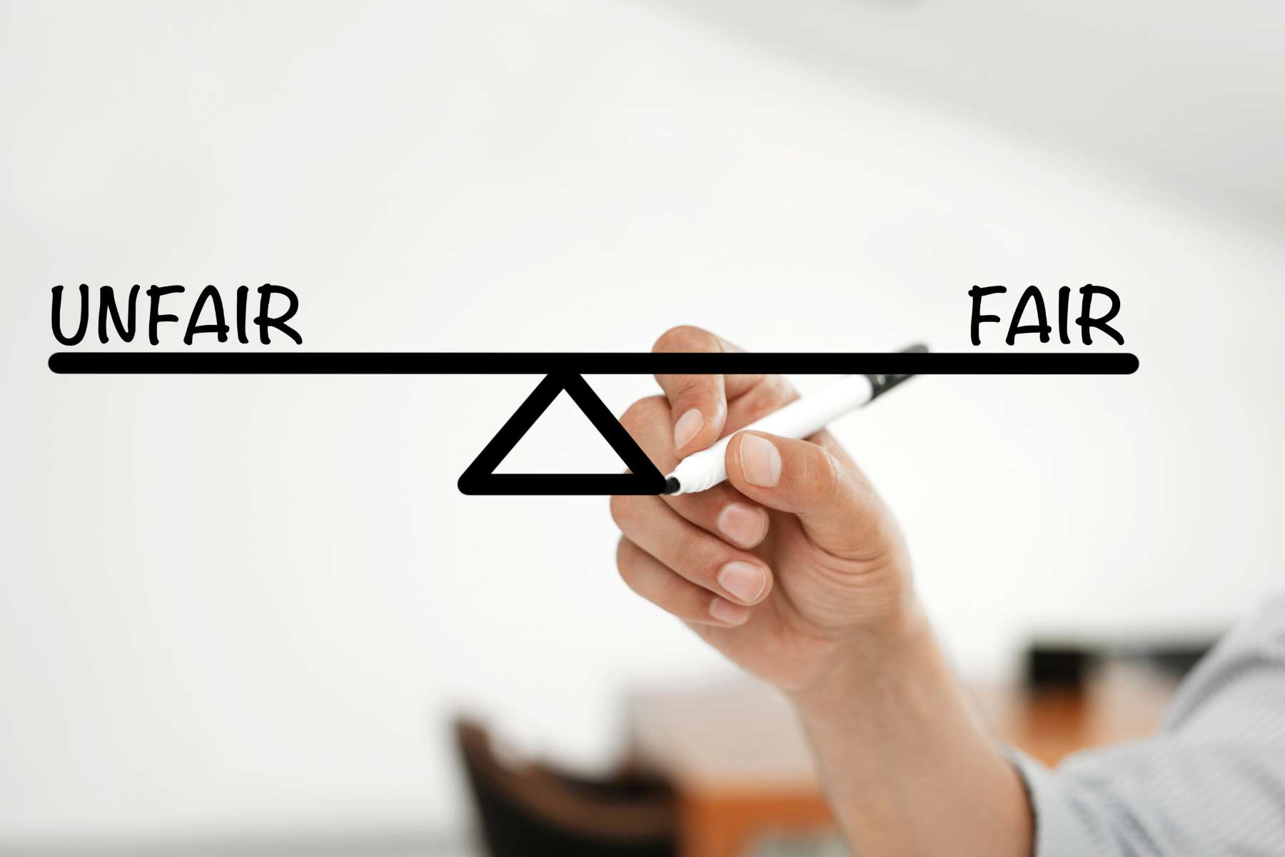 Eine Hand hält einen schwarzen Marker und malt damit eine vereinfachte Waage auf eine Glasscheibe, an deren einen Ende das Wort Unfair steht und an deren anderen Ende das Wort Fair.