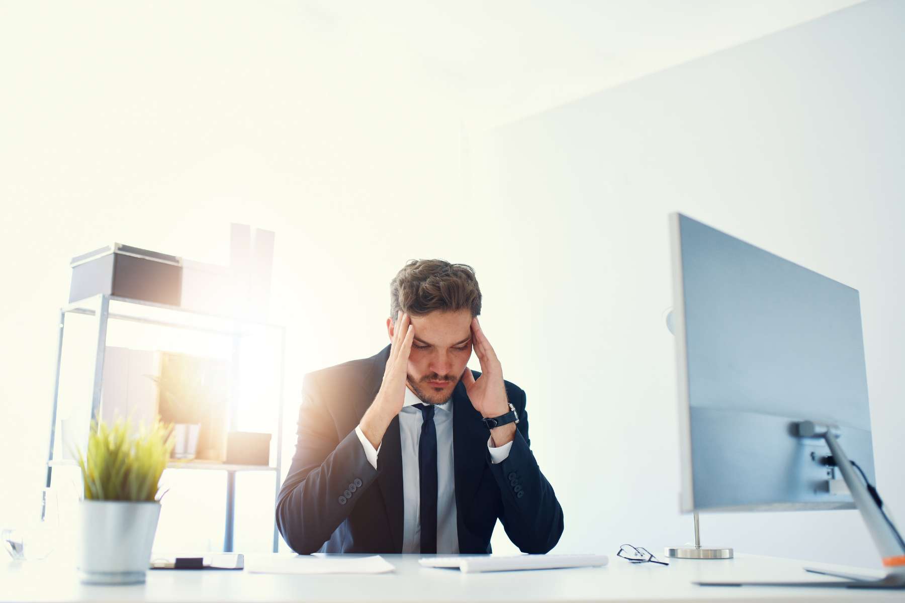 Ein erschöpft wirkender junger Mann im Anzug sitzt in einem Büro an einem Schreibtisch und hält sich mit geschlossenen Augen die flachen Hände an die Schläfen.