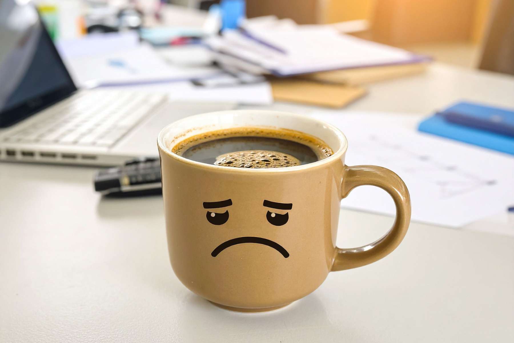 Eine gelbe Kaffeetasse, auf der ein unglückliches Gesicht abgebildet ist, steht auf einem vollen Schreibtisch in einem Büro, als Sinnbild zum Thema Return to Office.
