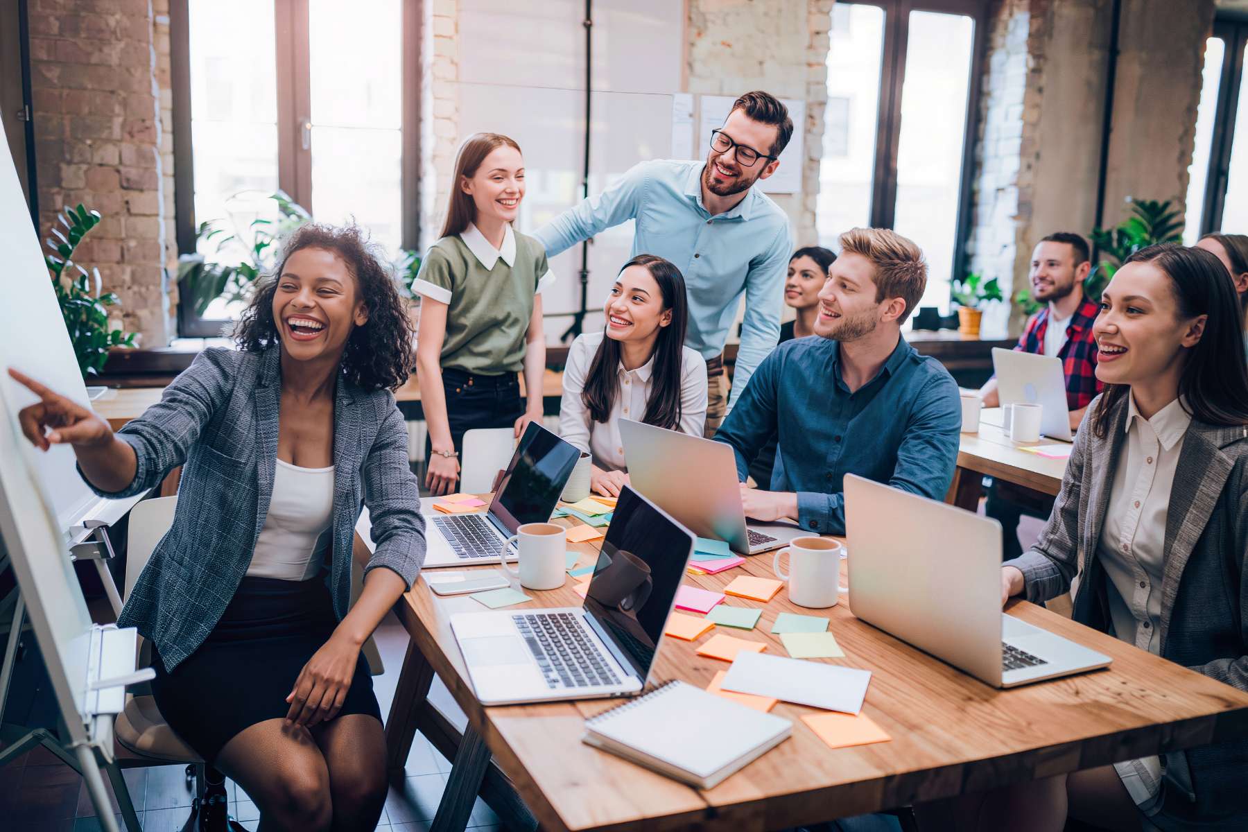 Eine Gruppe junger, diverser Büroangestellter sitzt lachend am Tisch, arbeitet zusammen und unterhält sich im Zuge einer Teambesprechung, als Sinnbild zum Thema Return to Office.