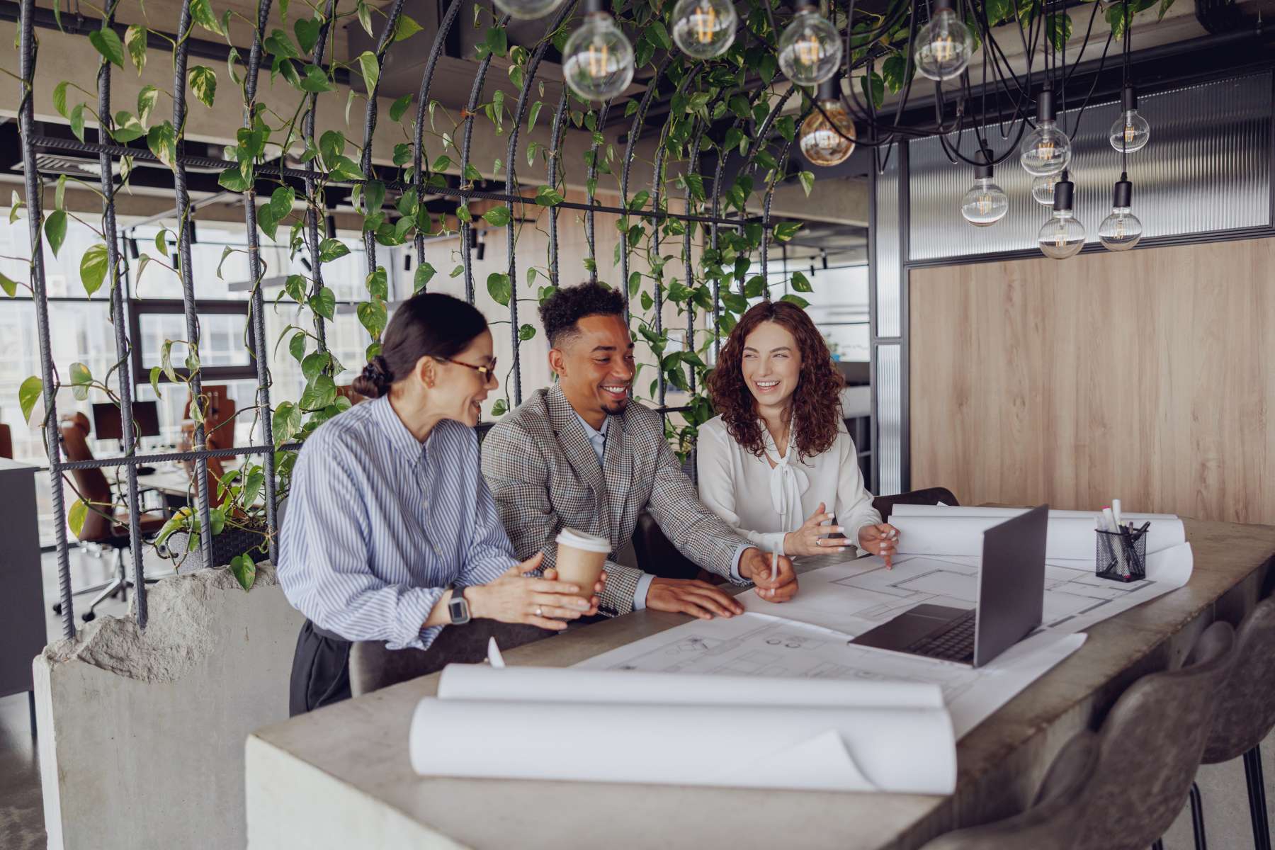 Drei Mitarbeiter sitzen in einem Büro an einem grauen Tisch aus Stein, arbeiten an einem Projekt und scherzen dabei, während man im Hintergrund ein biophiles Designelement in Form von Ranken sieht.