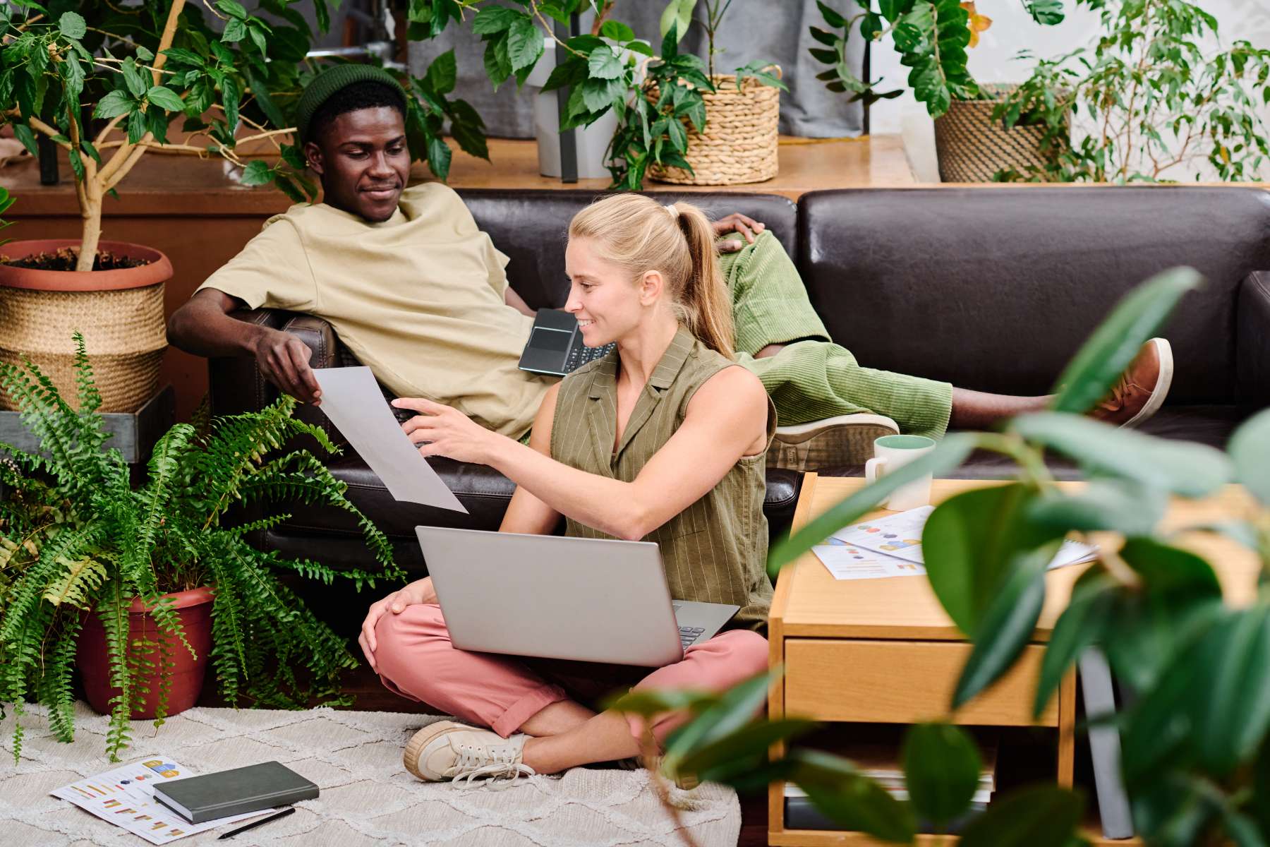 Eine weibliche Angestellte und ein männlicher Angestellter sitzen mit ihren Laptops in einer Loungeecke mit Pflanzen, flachem Holztisch und schwarzem Sofa und unterhalten sich angeregt.