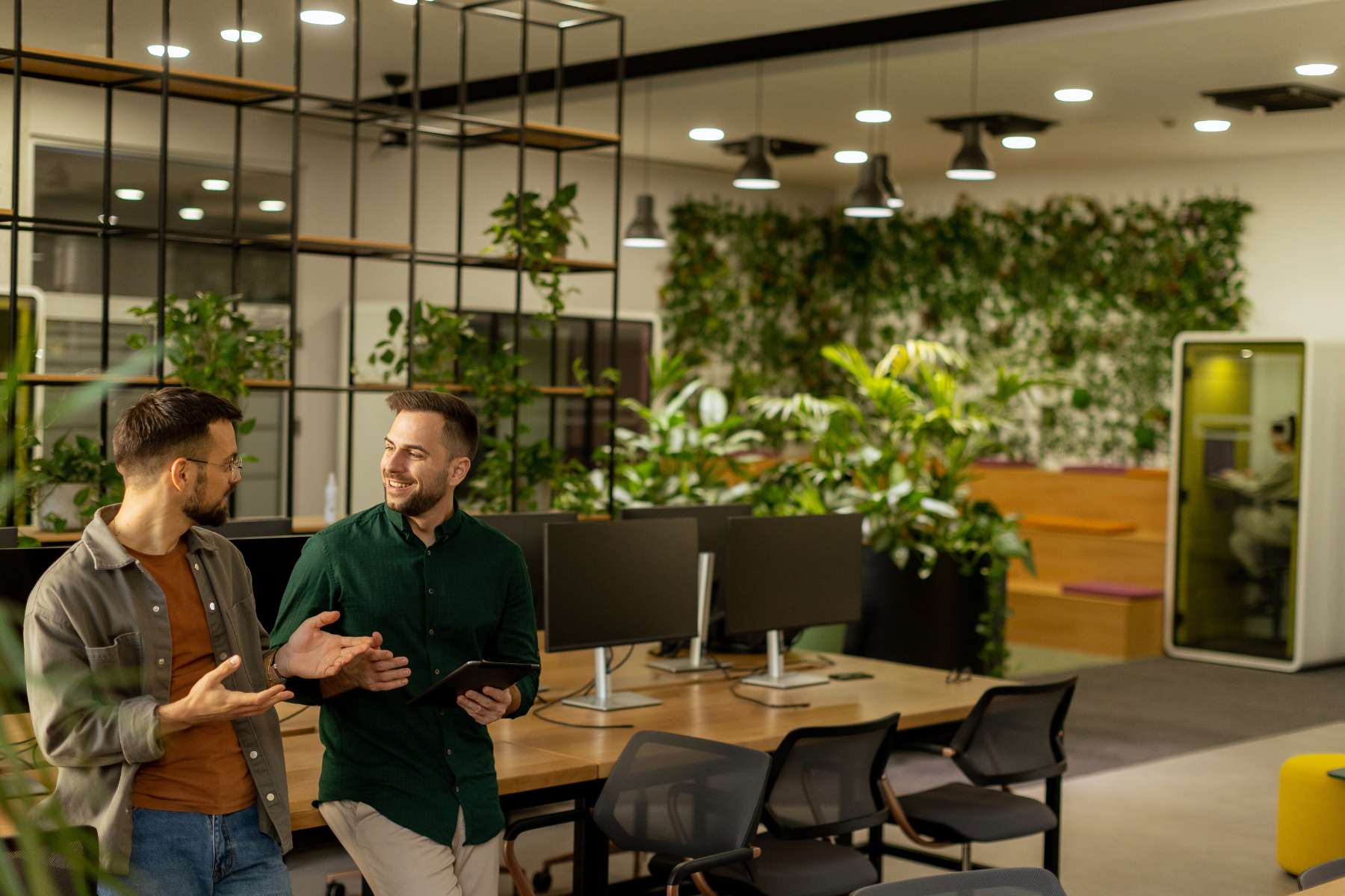 Zwei männliche Angestellte stehen in einem modernen Büro mit biophilem Design, lehnen an einem langen Holztisch mit schwarzen Monitoren und unterhalten sich angeregt.