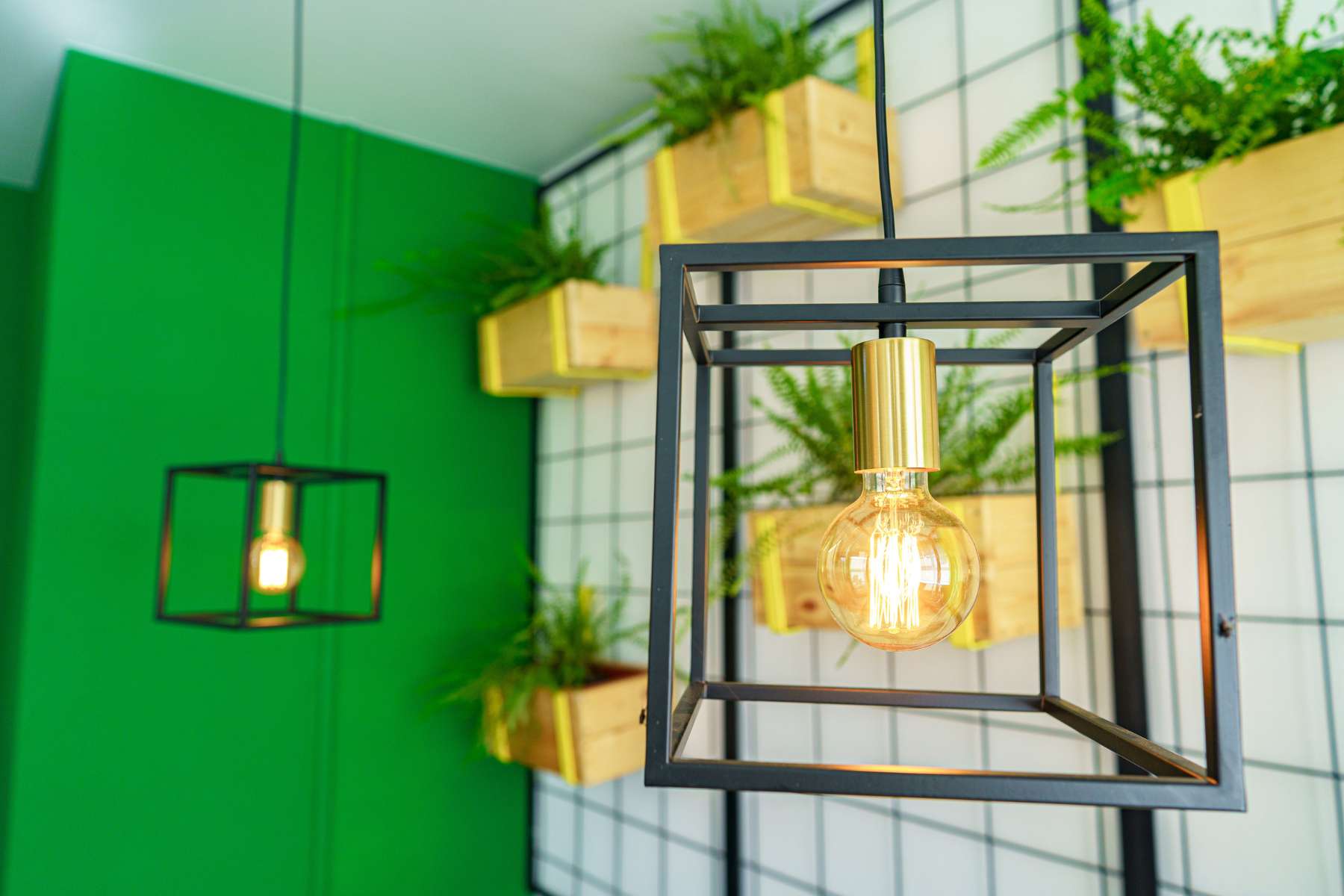 Hängelampe in Gold mit schwarzer Metallumrandung im Vordergrund, weitere Lampe und hängende Pflanzkästen aus Holz im Hintergrund als Beispiel für biophiles Design.