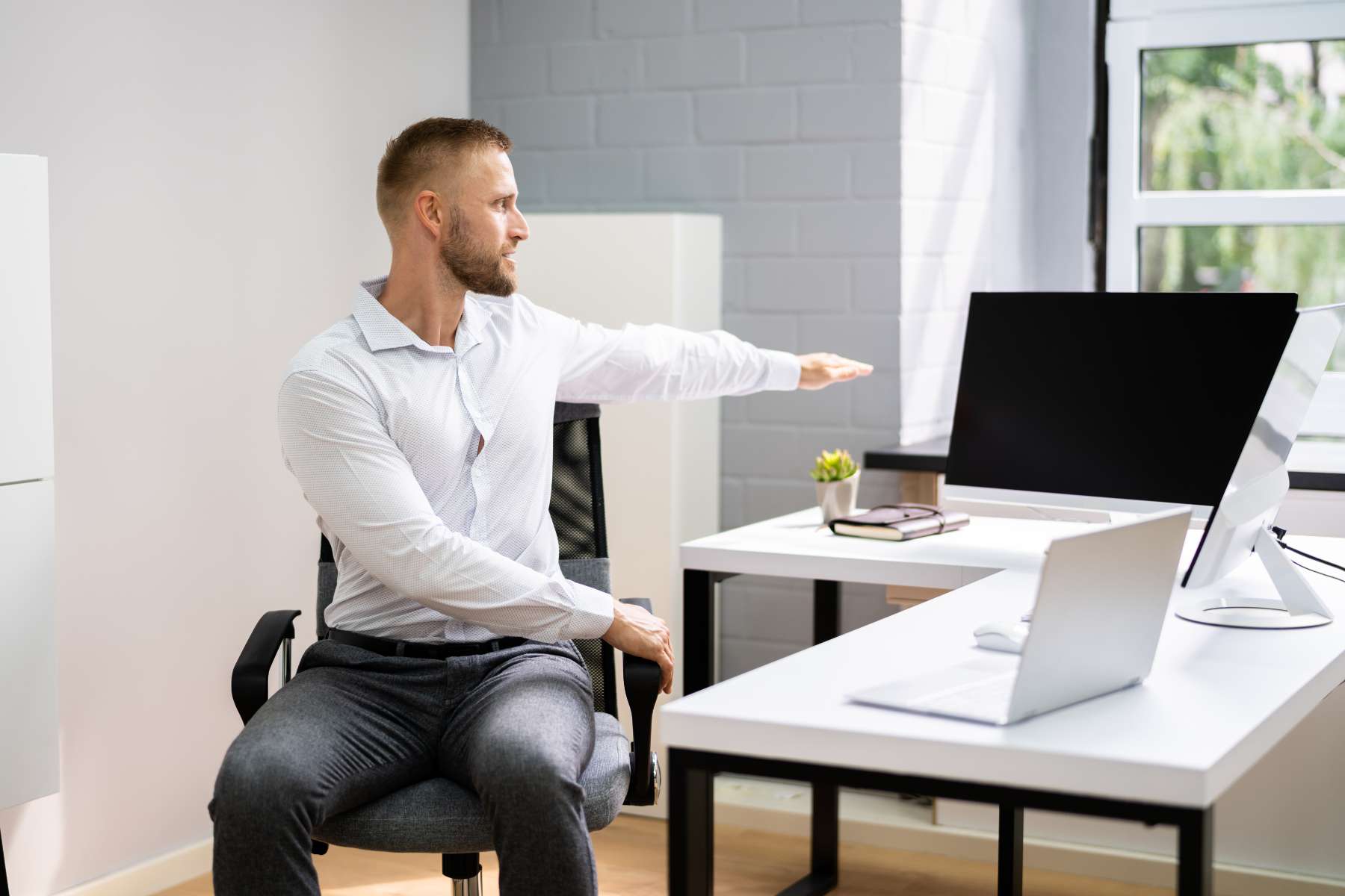 Ein junger Mann sitzt auf einem Drehstuhl vor einem Schreibtisch und macht eine Dehnübung als Maßnahme für Wellbeing im Büro.