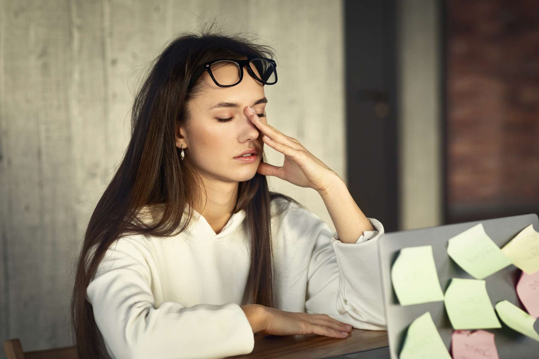 Eine gestresste junge Frau sitzt mit geschlossenen Augen am Schreibtisch vor einem Laptop und hält sich die Hand an die Stirn.