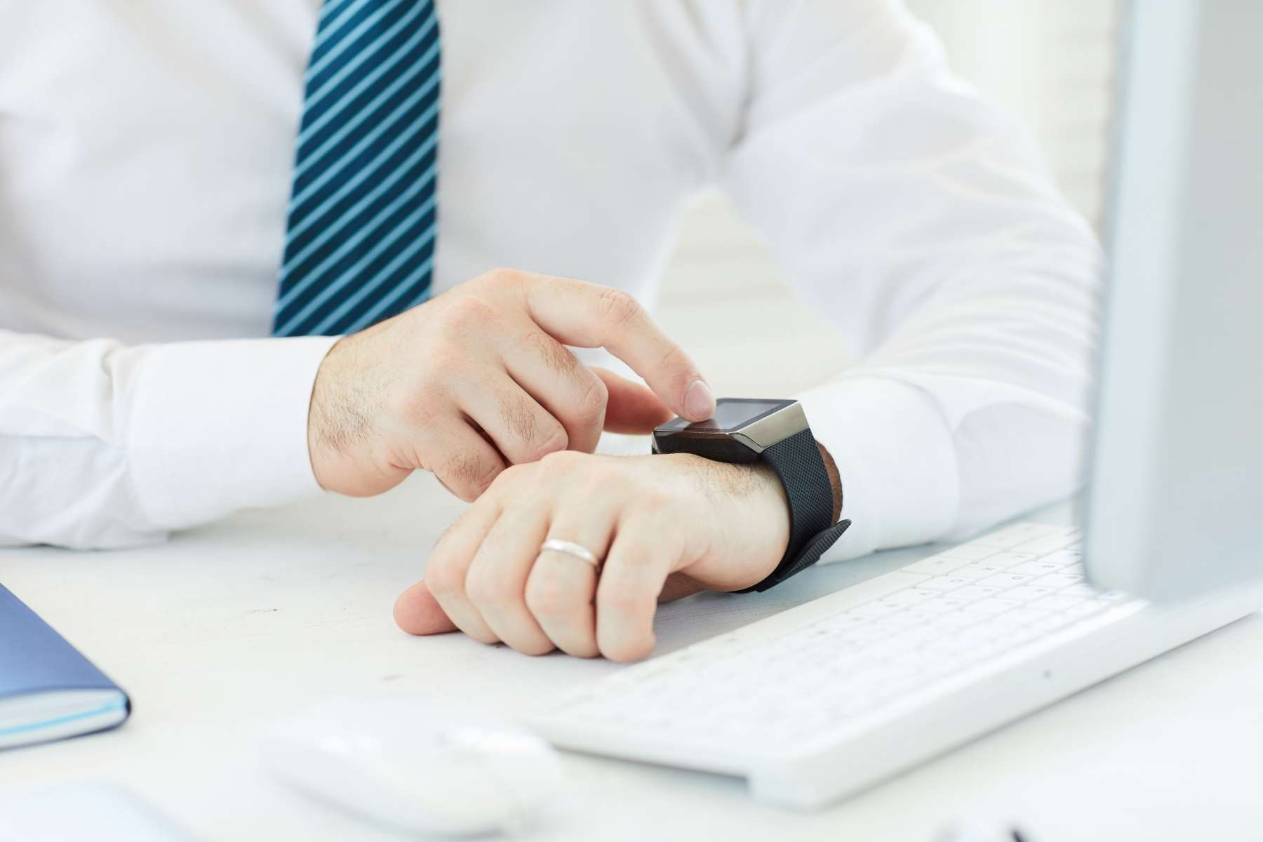 Ein Mann mit Krawatte und weißem Hemd sitzt an einem Schreibtisch vor einer Tastatur und schaut auf seine Smartwatch.